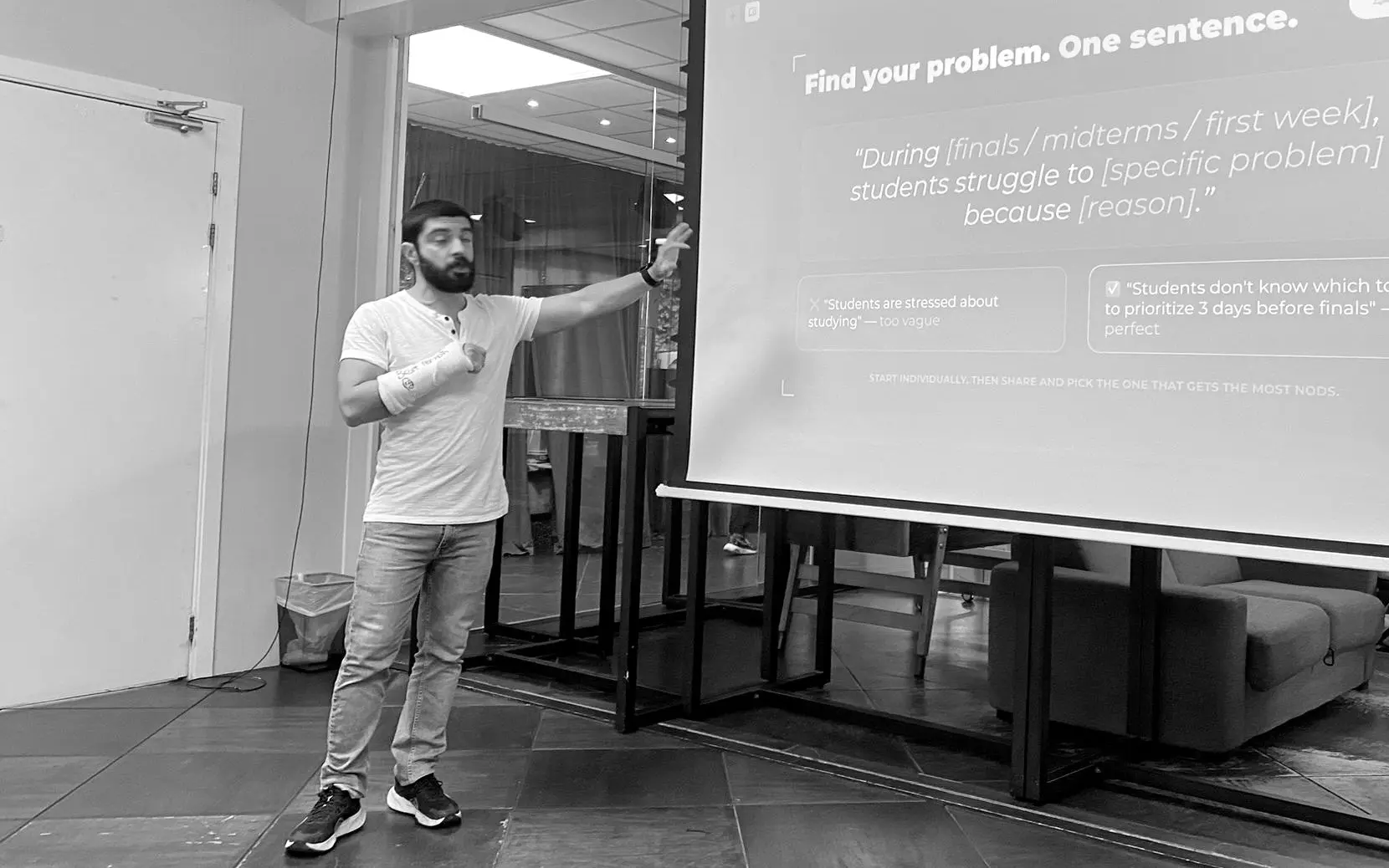Ömer at the front of the room, pointing at a projected slide that reads Find your problem, one sentence, with the fill-in-the-blanks problem format under it