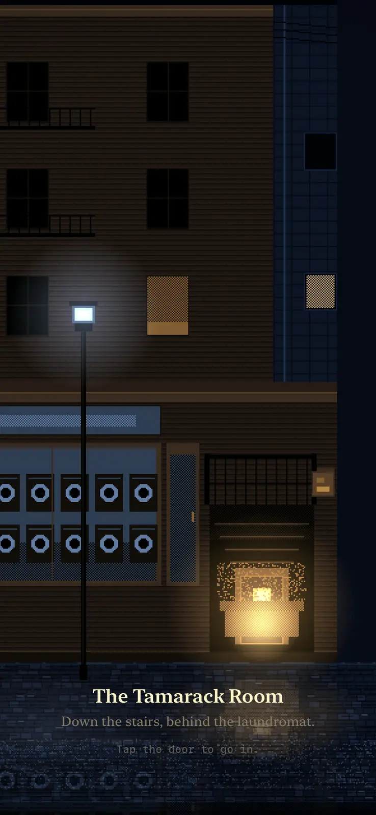 A pixel art street at night in New York. A brick apartment block with dark windows, a laundromat with two rows of round machine doors, a lamp post, and beside it a lit doorway at the foot of a short stair. The title reads The Tamarack Room, then Down the stairs, behind the laundromat, then Tap the door to go in. Wet paving reflects the light.