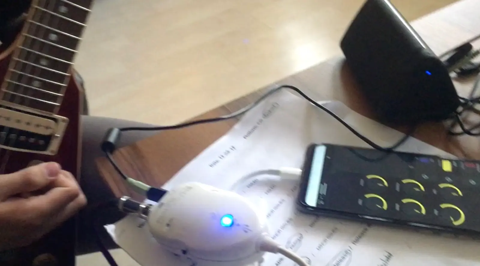 A test on a desk. A hand on the strings of an electric guitar at the left, its cable running into a small white interface lit blue, and on from there to a phone lying on a sheet of notes, open on the studio with its knobs lit yellow. A speaker stands behind.