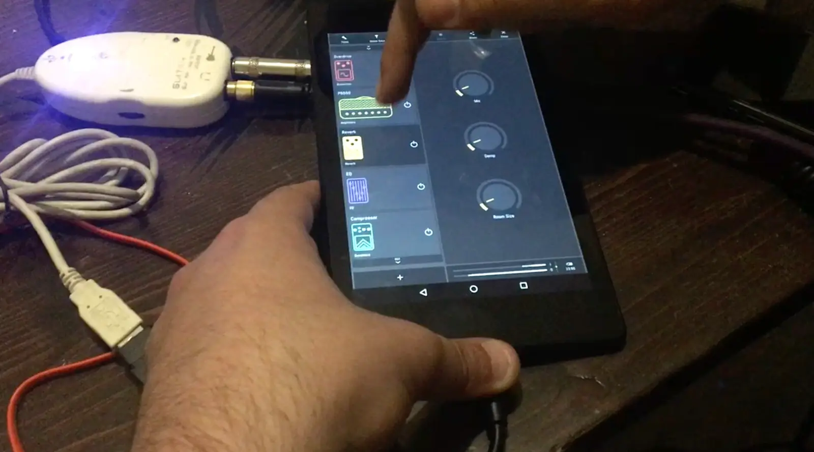 A tablet on a dark table, plugged into a white guitar interface at the top left. A hand points at the chain in the studio's left column.