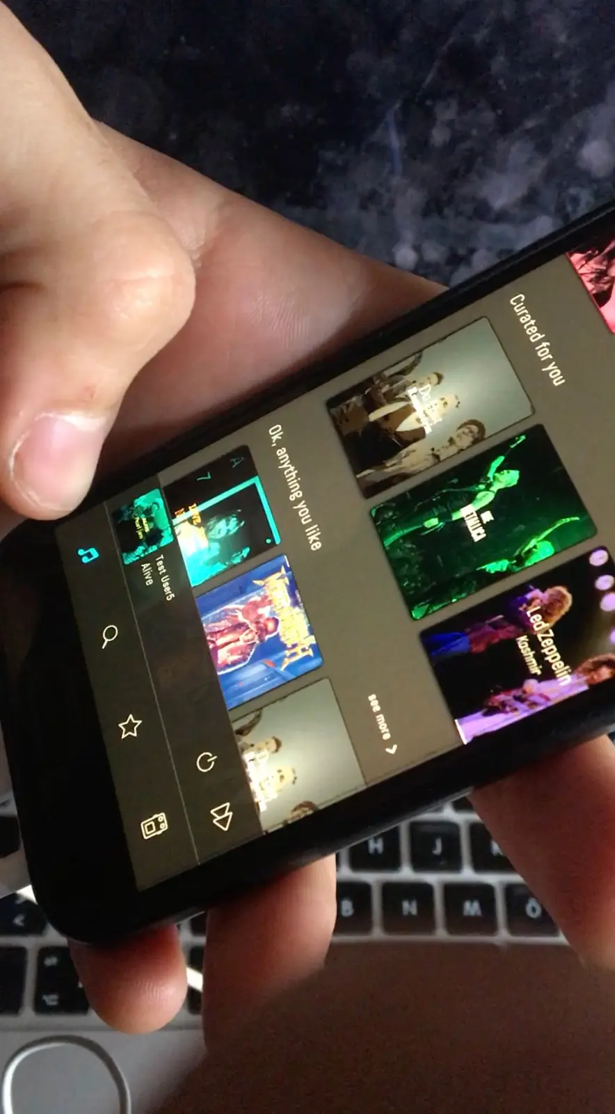 A phone held sideways in two hands over a laptop, open on the home feed, a column of tone cards over band photographs.